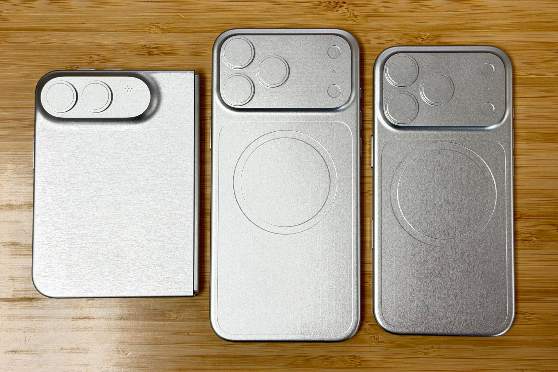 Three silver metal mockups, including an iPhone Fold and other smartphones, rest on a wooden surface. Each mockup features precise camera cutouts and a distinctive circular design on the back.