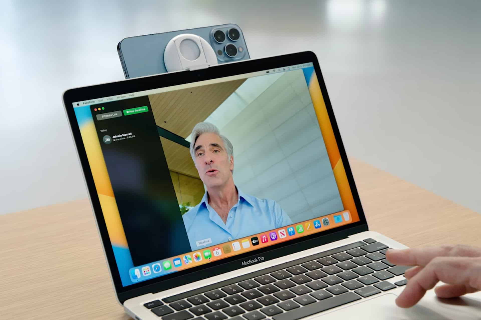 A laptop displays a video call with a man speaking; an iPhone, using Mac Continuity Camera, is mounted on top of the laptop as a webcam. A hand points at the laptop trackpad.