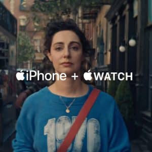 A woman with curly hair, wearing a blue "1970" sweatshirt and a red strap, walks down a city street. The text "iPhone + Apple Watch: Apple Health pairing" with Apple logos appears across the image.