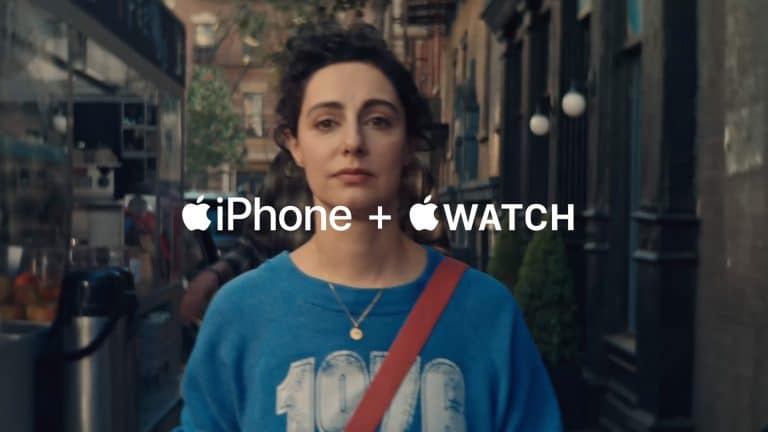 A woman with curly hair, wearing a blue "1970" sweatshirt and a red strap, walks down a city street. The text "iPhone + Apple Watch: Apple Health pairing" with Apple logos appears across the image.