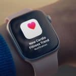 A smartwatch on a person's wrist, paired with Apple Health, displays a notification with a pink heart icon and the message, "New Cardio Fitness Trend Notification." The person is wearing a blue sleeve.