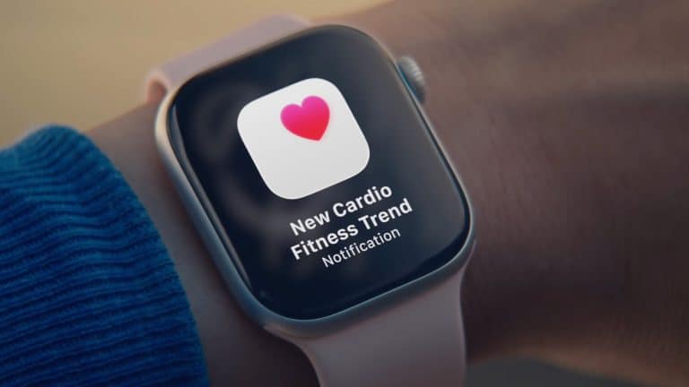 A smartwatch on a person's wrist, paired with Apple Health, displays a notification with a pink heart icon and the message, "New Cardio Fitness Trend Notification." The person is wearing a blue sleeve.