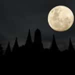 A bright full moon shines in a dark, cloudy sky above the silhouette of pointed temple structures and trees at night, captured perfectly in an iPhone moon shot.
