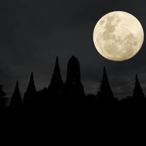 A bright full moon shines in a dark, cloudy sky above the silhouette of pointed temple structures and trees at night, captured perfectly in an iPhone moon shot.