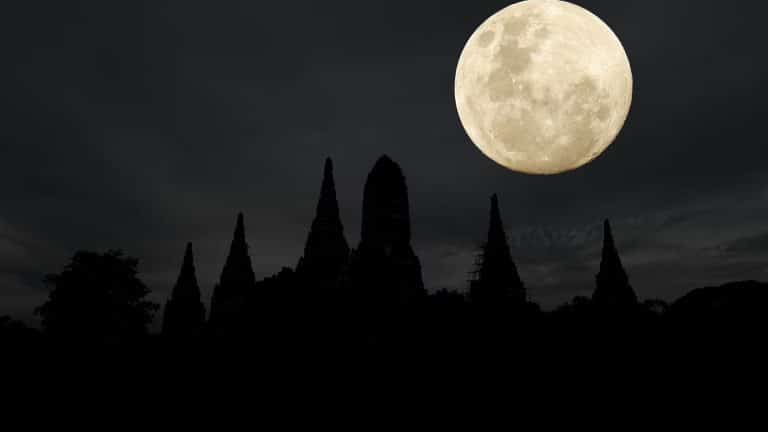 A bright full moon shines in a dark, cloudy sky above the silhouette of pointed temple structures and trees at night, captured perfectly in an iPhone moon shot.