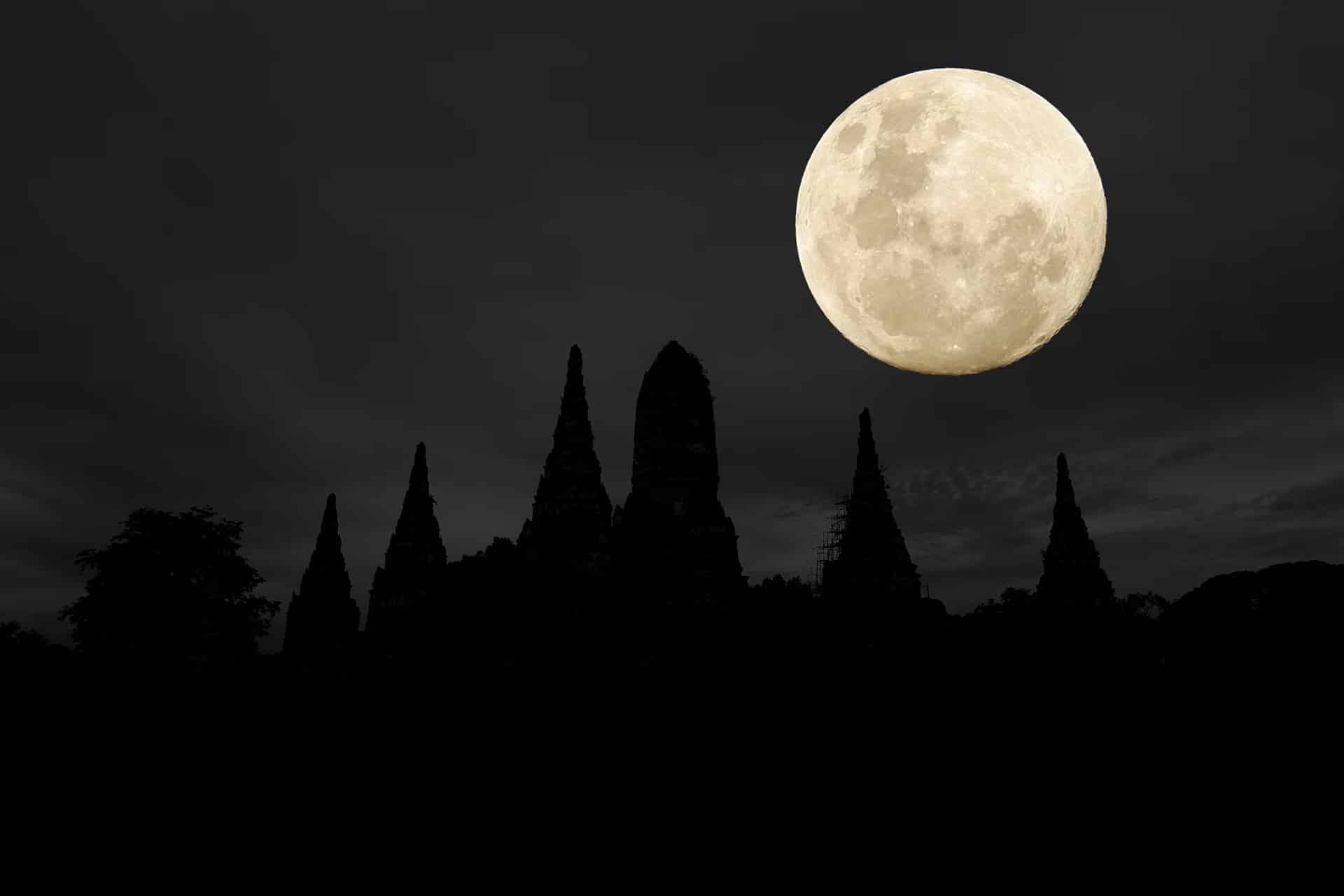 A bright full moon shines in a dark, cloudy sky above the silhouette of pointed temple structures and trees at night, captured perfectly in an iPhone moon shot.