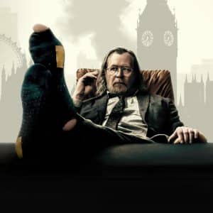 A man in a suit sits back in a chair with his feet up on a desk, wearing dark socks. He looks relaxed, holding glasses in one hand. The London Eye and Big Ben rise behind him—a scene fit for celebrating BAFTA wins.