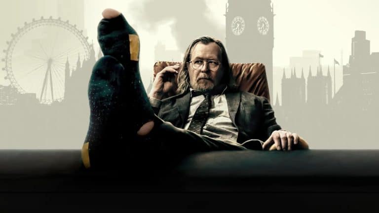 A man in a suit sits back in a chair with his feet up on a desk, wearing dark socks. He looks relaxed, holding glasses in one hand. The London Eye and Big Ben rise behind him—a scene fit for celebrating BAFTA wins.