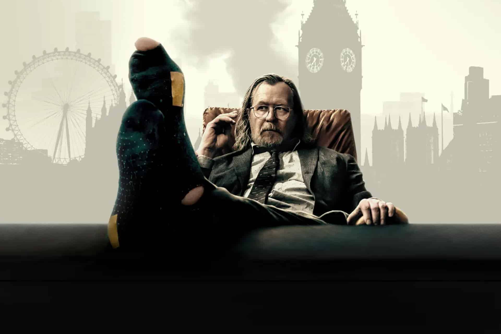 A man in a suit sits back in a chair with his feet up on a desk, wearing dark socks. He looks relaxed, holding glasses in one hand. The London Eye and Big Ben rise behind him—a scene fit for celebrating BAFTA wins.