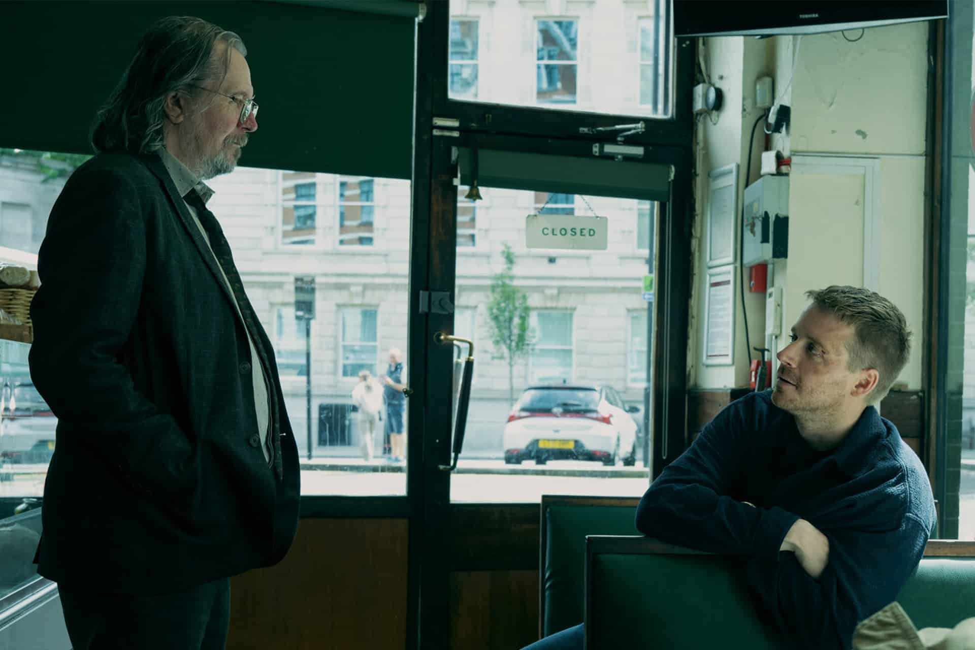 Two men are having a conversation inside a cafe. One man with long hair and glasses stands, while the other, younger man sits on a bench with his arms crossed. Through the glass door marked “Closed,” a parked car and posters about BAFTA wins are visible.