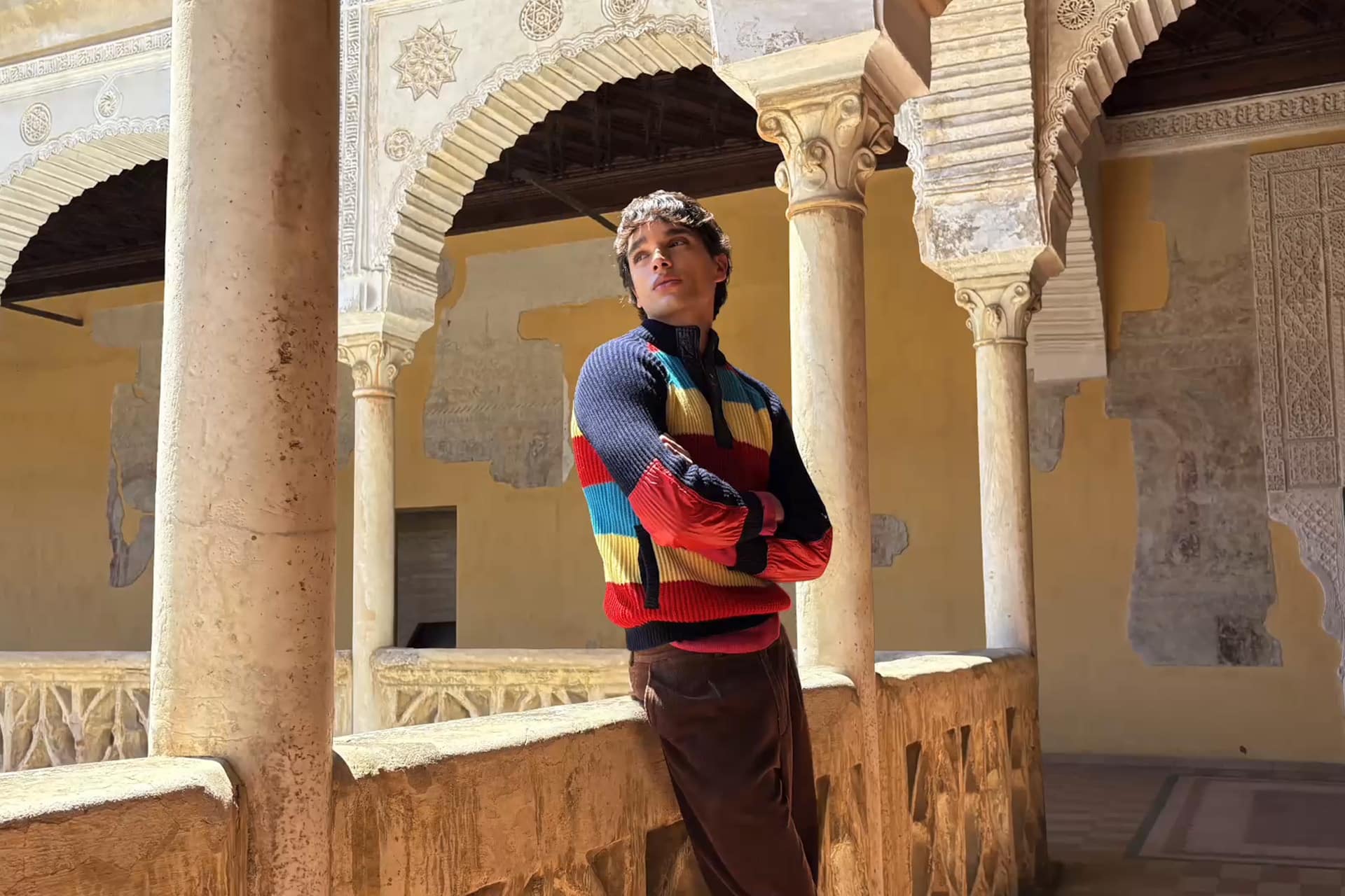 A person in a colorful sweater leans against a stone railing in a historic building with ornate arches and columns, thoughtfully observing the spatial scenes around them as sunlight highlights the intricate architectural details.