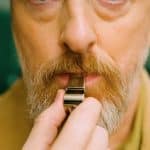 A close-up of a man’s face with light skin, a mustache, and a beard as he holds a silver whistle to his mouth, preparing to blow it—capturing the anticipation just before "After the Whistle." The background is blurred green.