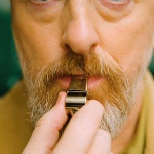 A close-up of a man’s face with light skin, a mustache, and a beard as he holds a silver whistle to his mouth, preparing to blow it—capturing the anticipation just before "After the Whistle." The background is blurred green.