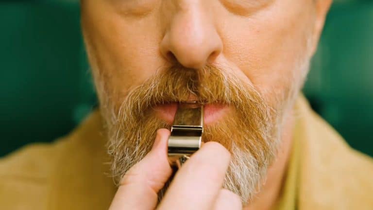 A close-up of a man’s face with light skin, a mustache, and a beard as he holds a silver whistle to his mouth, preparing to blow it—capturing the anticipation just before "After the Whistle." The background is blurred green.
