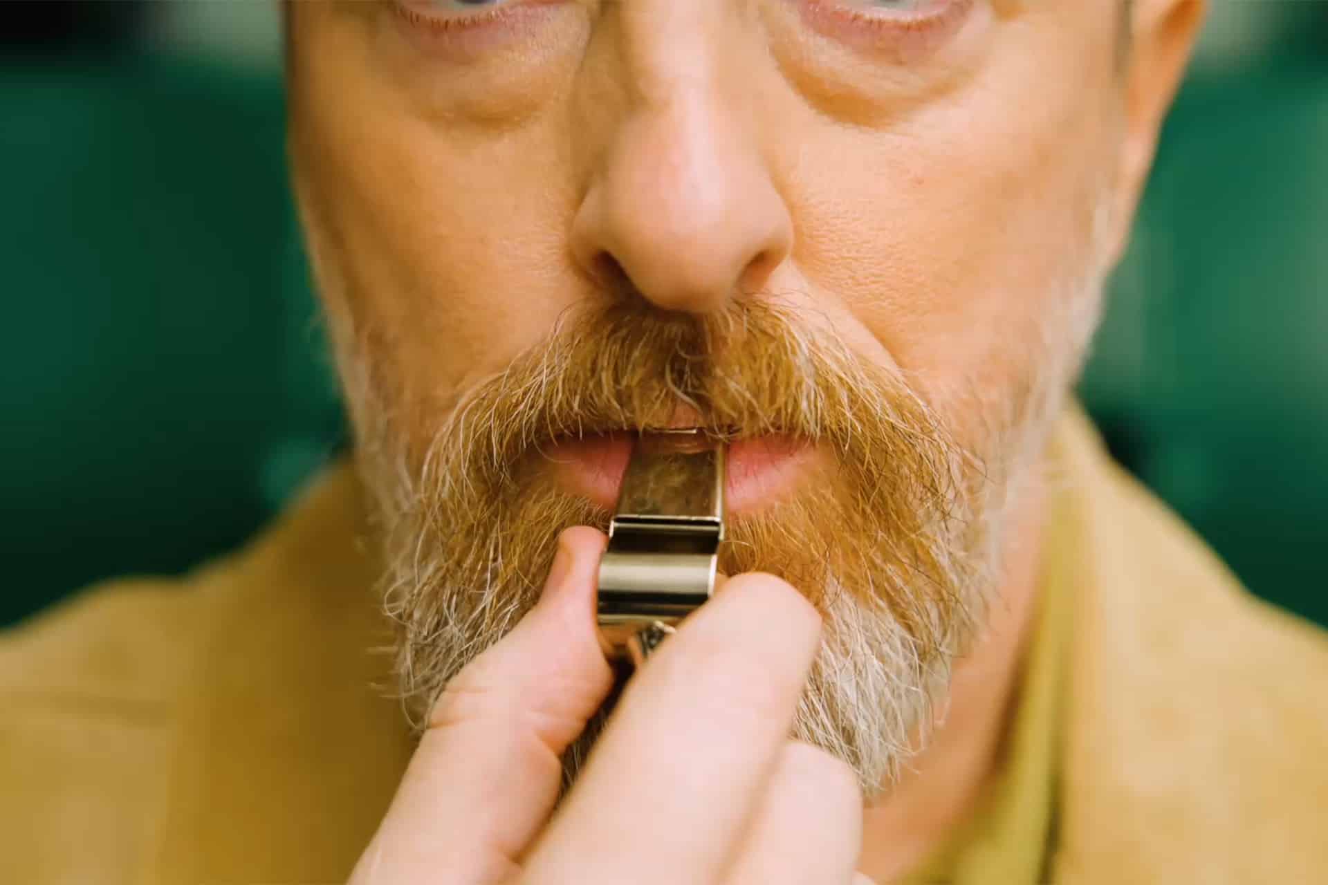 A close-up of a man’s face with light skin, a mustache, and a beard as he holds a silver whistle to his mouth, preparing to blow it—capturing the anticipation just before "After the Whistle." The background is blurred green.