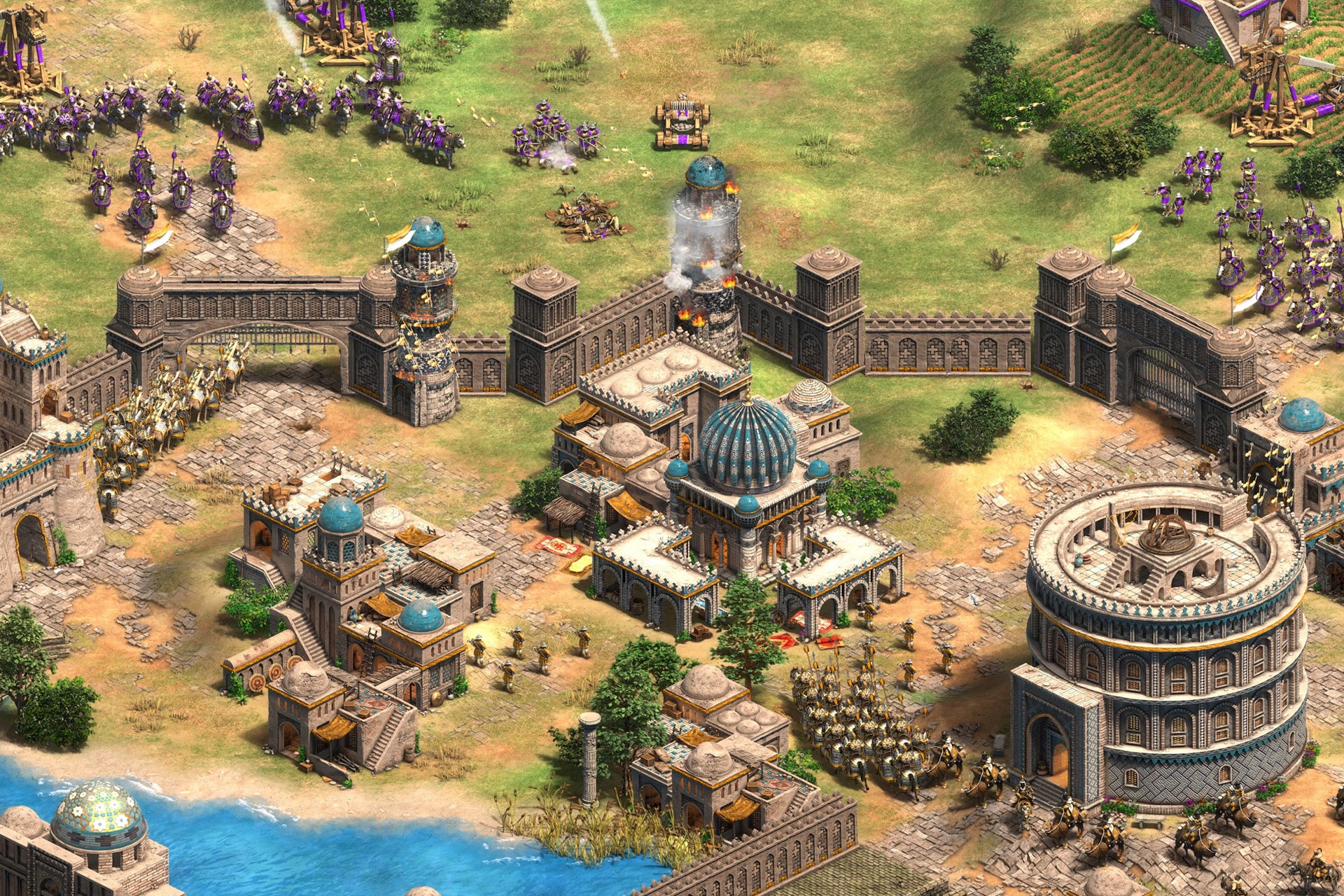A detailed isometric view of a fortified medieval city from Age of Empires II Mac, with stone walls, towers, domed buildings, cavalry, and infantry units. The city is under attack, smoke rising from a central building—a classic scene in macOS gaming.