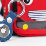 A close-up of a blue Apple AirTag keychain attached to a red bag with a cartoon face and yellow circles, resembling a toy vehicle—perfect for using Find My for kids to keep track of their belongings.