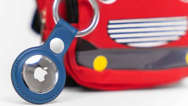 A close-up of a blue Apple AirTag keychain attached to a red bag with a cartoon face and yellow circles, resembling a toy vehicle—perfect for using Find My for kids to keep track of their belongings.