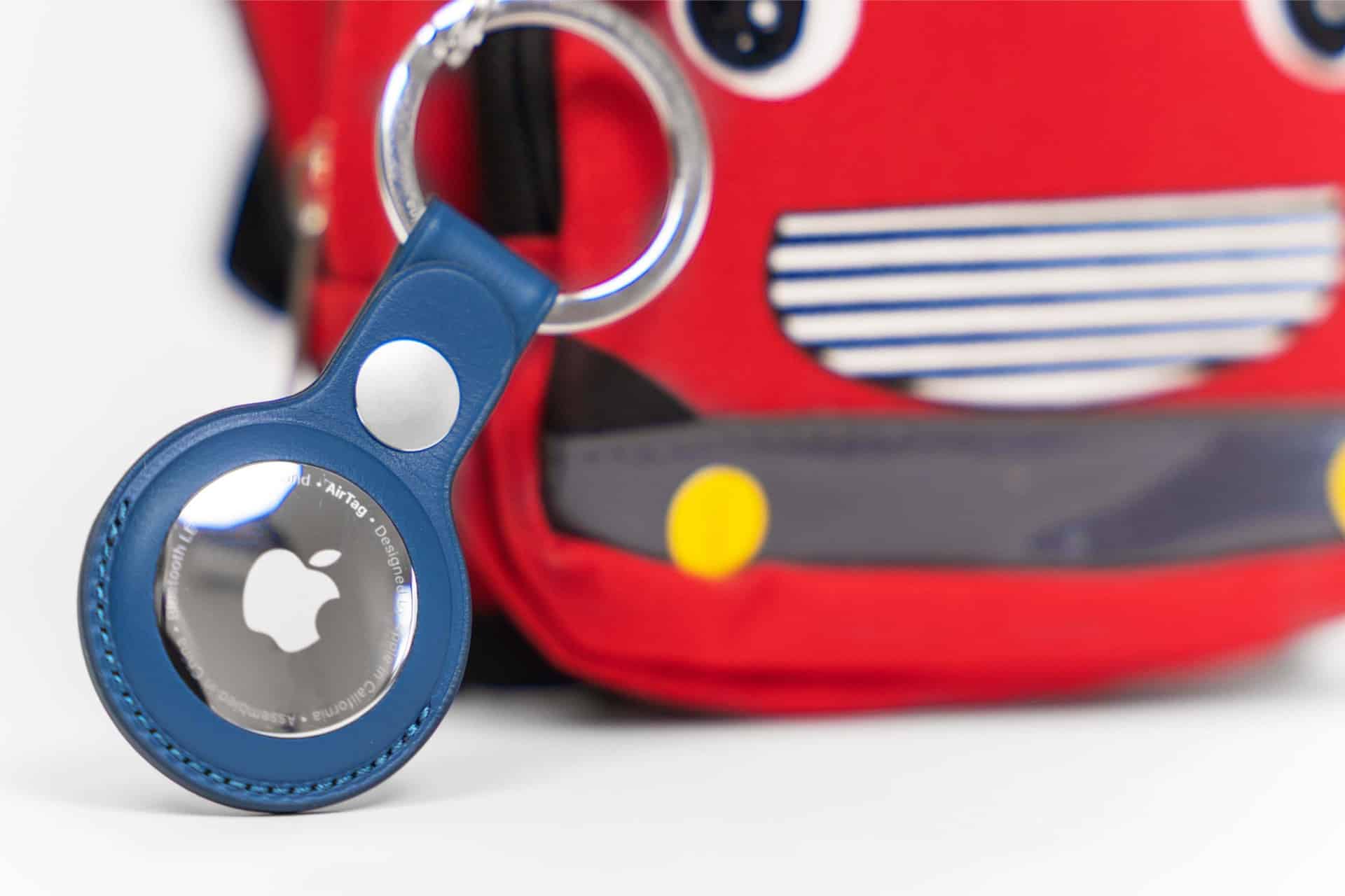 A close-up of a blue Apple AirTag keychain attached to a red bag with a cartoon face and yellow circles, resembling a toy vehicle—perfect for using Find My for kids to keep track of their belongings.