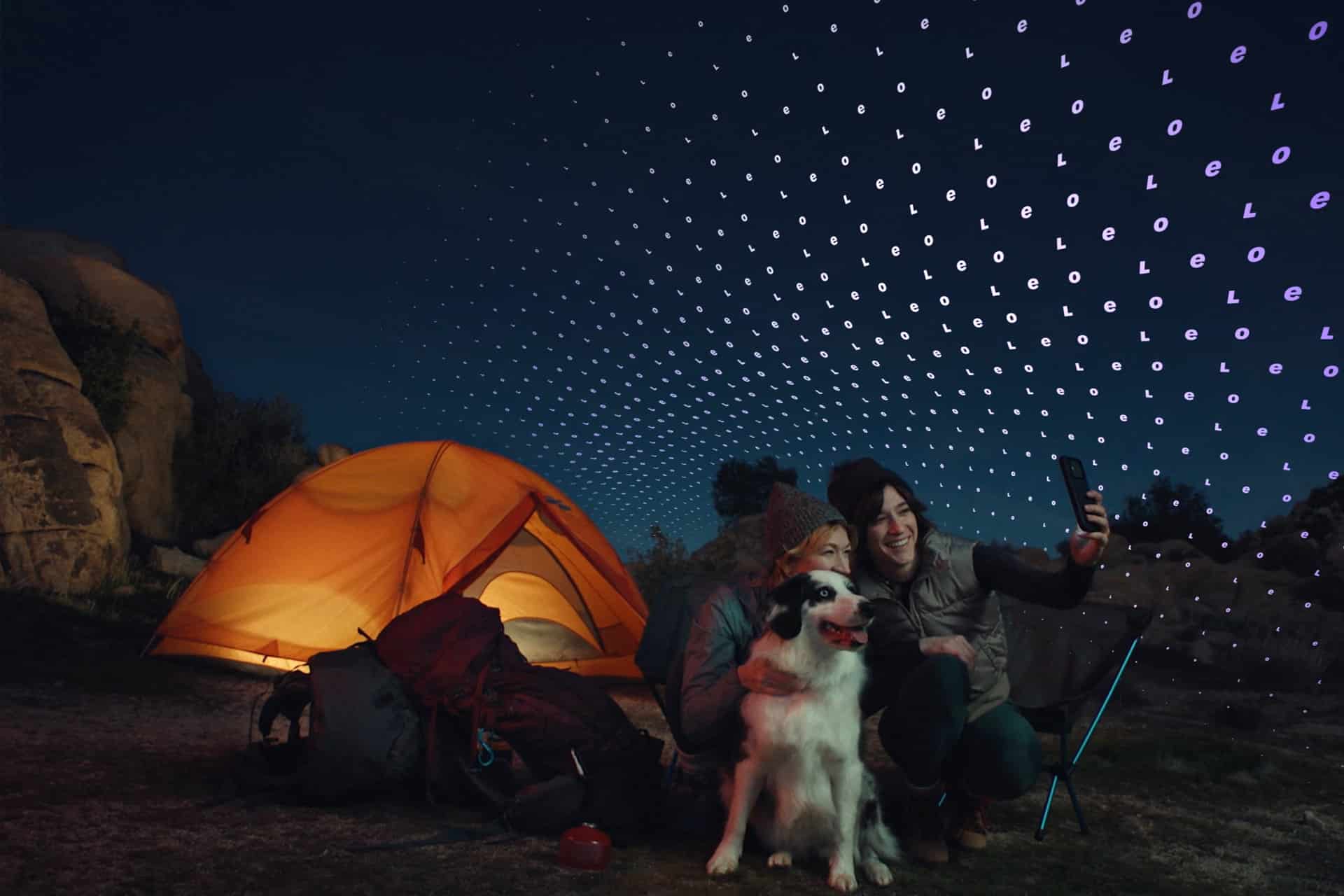 Two people and a dog pose for a selfie beside a glowing orange tent at night; above them, a digital grid with binary code suggests an Amazon Globalstar satellite internet connection linking their remote adventure.