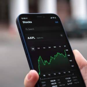 A person holds a smartphone displaying the Stocks app, showing Apple Inc. (AAPL) stock performance with a fluctuating green line graph—potentially influenced by recent Apple R&D spending—against a black background.