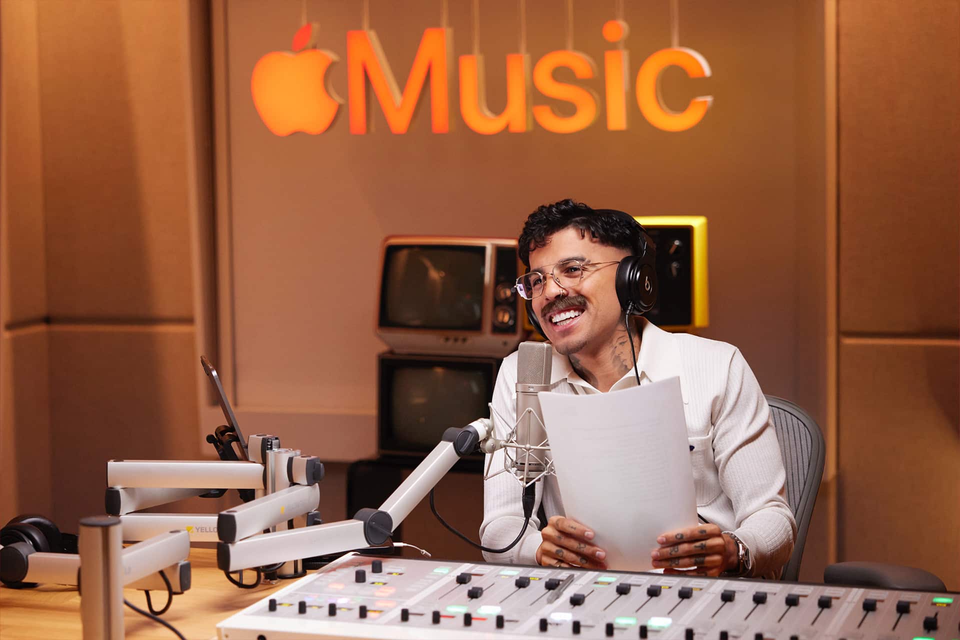 A man wearing headphones and holding papers speaks into a microphone in a recording studio with an Apple Music Radio sign glowing in the background. Old TVs and audio equipment are visible behind him.