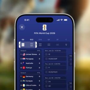Close-up of a smartphone using Live Activities in the Apple Sports app to display the FIFA World Cup 2026 schedule, highlighting group stage matches for the United States, Paraguay, Australia, and Turkey. A blurred soccer field and players fill the background.