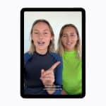 Two people sit side by side, facing the camera on a tablet screen using Apple accessibility features. One gestures while speaking, and subtitles read, "Seagulls are very territorial apparently." Both wear rash guards, one black and one neon green.