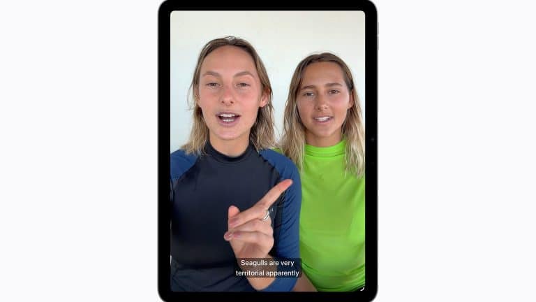 Two people sit side by side, facing the camera on a tablet screen using Apple accessibility features. One gestures while speaking, and subtitles read, "Seagulls are very territorial apparently." Both wear rash guards, one black and one neon green.
