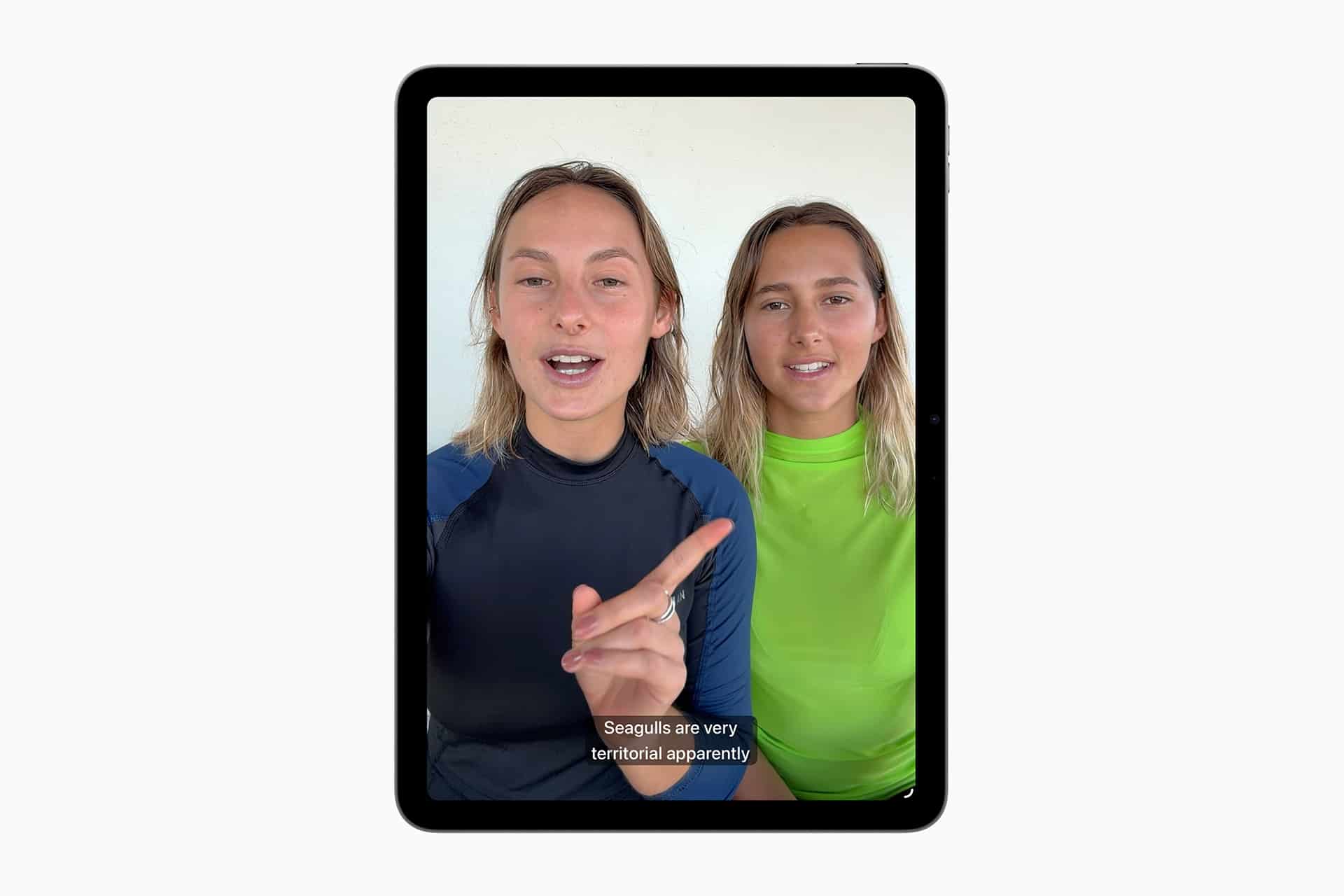 Two people sit side by side, facing the camera on a tablet screen using Apple accessibility features. One gestures while speaking, and subtitles read, "Seagulls are very territorial apparently." Both wear rash guards, one black and one neon green.