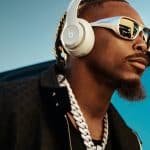 A man wearing white Beats audio headphones, reflective sunglasses, a thick silver chain, and a black jacket stands outdoors with a blue sky in the background.