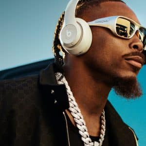 A man wearing white Beats audio headphones, reflective sunglasses, a thick silver chain, and a black jacket stands outdoors with a blue sky in the background.