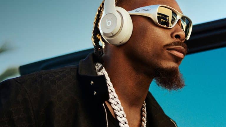 A man wearing white Beats audio headphones, reflective sunglasses, a thick silver chain, and a black jacket stands outdoors with a blue sky in the background.