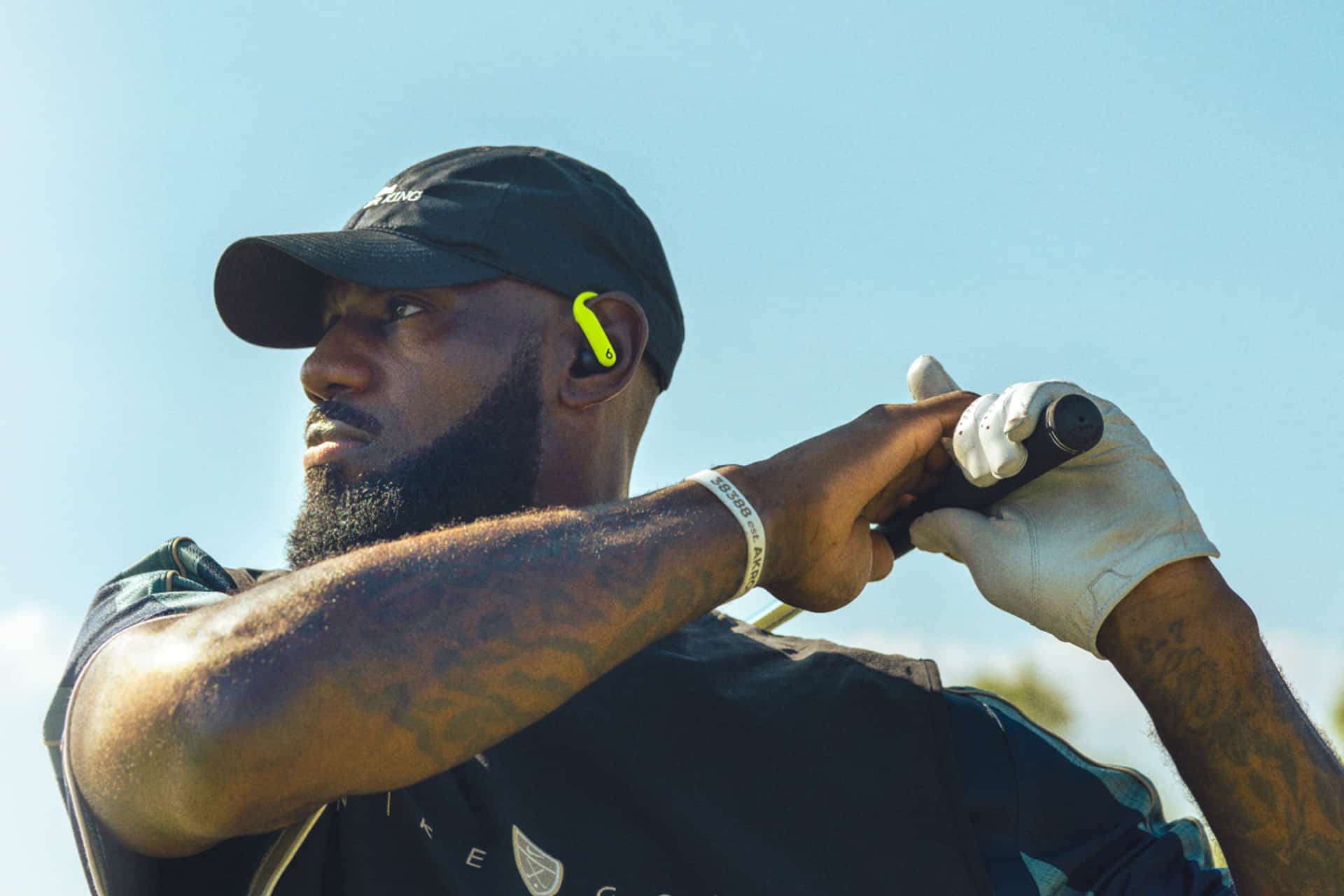 A man wearing a black cap and yellow Beats audio wireless earbud holds a baseball bat over his shoulder, looking to the side. He is wearing a white glove and has visible tattoos on his arm, standing outdoors under a clear blue sky.