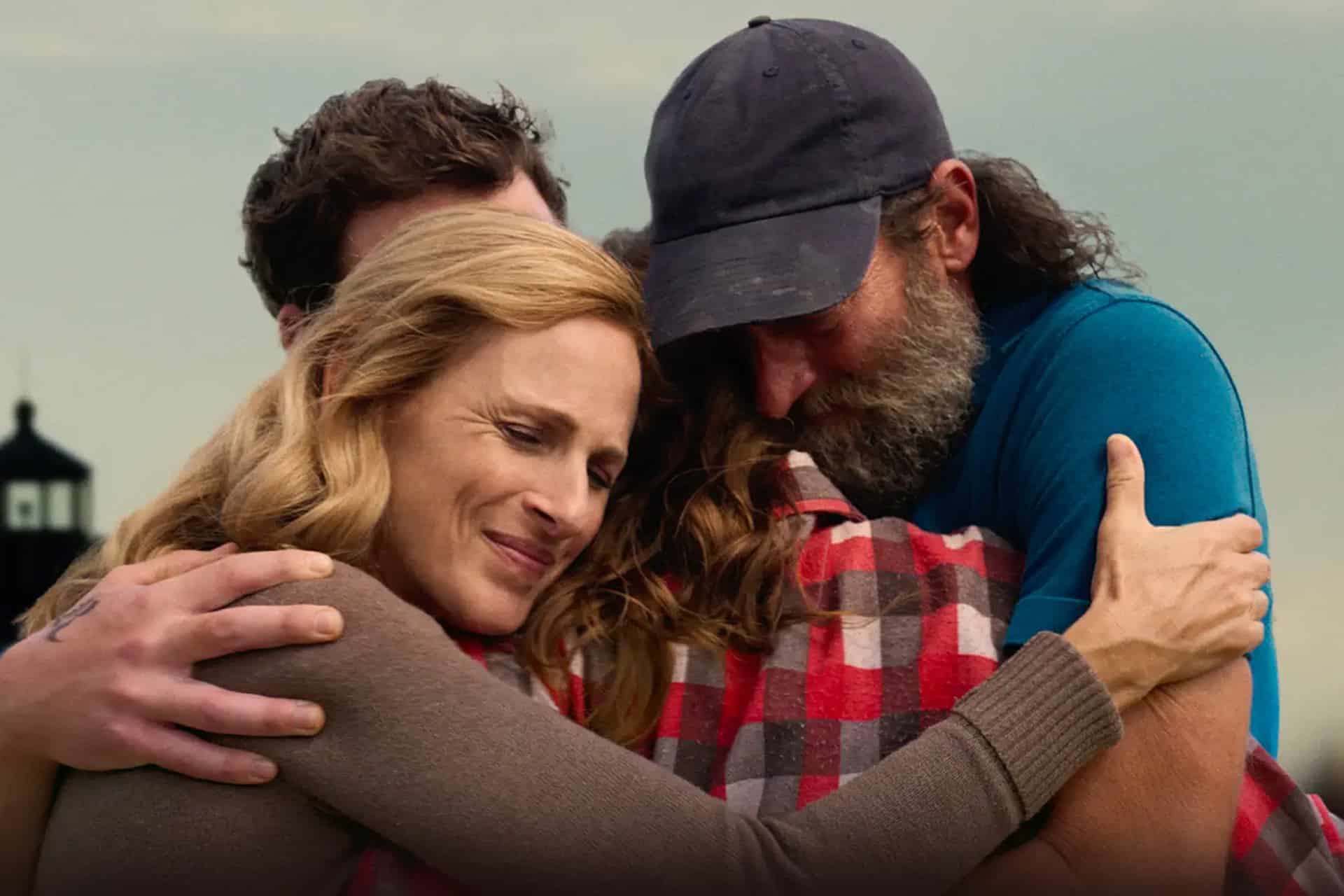 Three people embrace tightly outdoors in an emotional, heartfelt moment. A blonde woman smiles with her eyes closed as a bearded man in a baseball cap hugs someone in a red plaid shirt—capturing the warmth of an Apple TV weekend reunion.