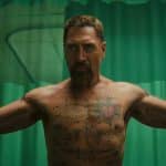 A muscular, bearded man with short hair stands shirtless, arms outstretched and tattoos on display, evoking the intensity of a Cape Fear thriller. He is in a room with green curtains and medical equipment visible in the background.