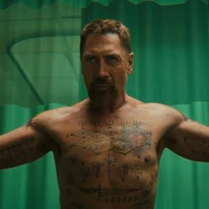A muscular, bearded man with short hair stands shirtless, arms outstretched and tattoos on display, evoking the intensity of a Cape Fear thriller. He is in a room with green curtains and medical equipment visible in the background.