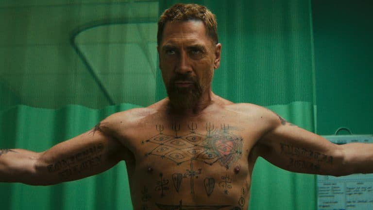 A muscular, bearded man with short hair stands shirtless, arms outstretched and tattoos on display, evoking the intensity of a Cape Fear thriller. He is in a room with green curtains and medical equipment visible in the background.
