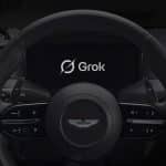 A car steering wheel with the Aston Martin logo, dashboard controls, and a central display screen showing "Grok Voice" with its logo. An Apple logo appears in the lower right corner.