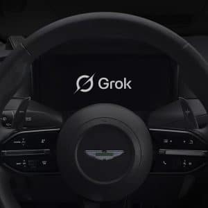 A car steering wheel with the Aston Martin logo, dashboard controls, and a central display screen showing "Grok Voice" with its logo. An Apple logo appears in the lower right corner.