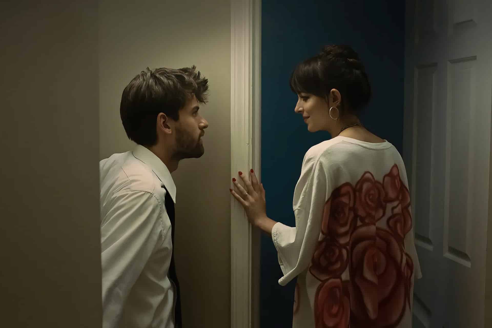 A man in a white shirt and tie leans against a doorway, talking with a woman in a white top with red rose designs, in the moody lighting of a hallway—just like a scene from an Apple TV movie.