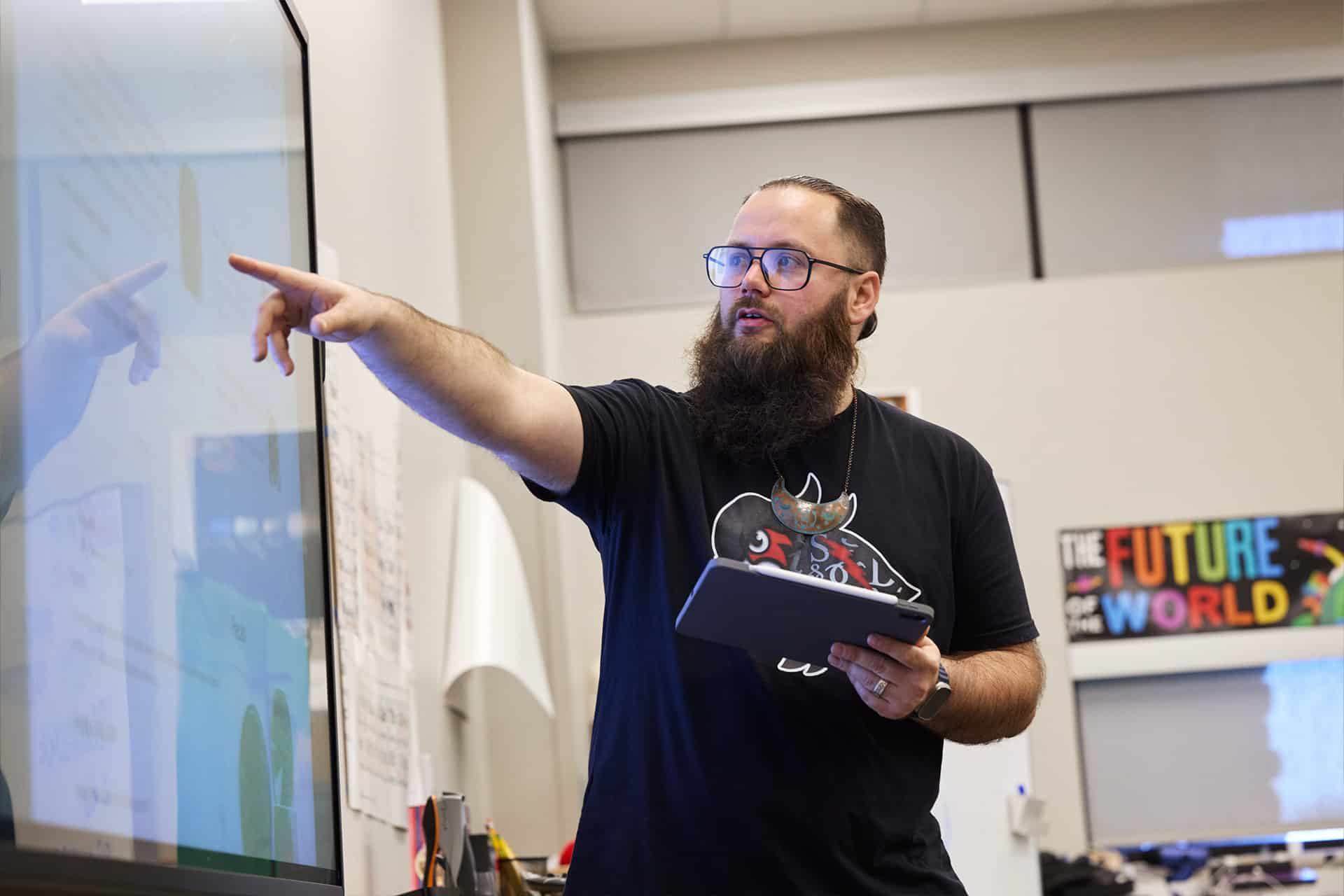 A bearded man wearing glasses and a black t-shirt points at a large touchscreen while holding an iPad displaying Cherokee language content. He is standing in a classroom with a colorful "The Future World" poster in the background.