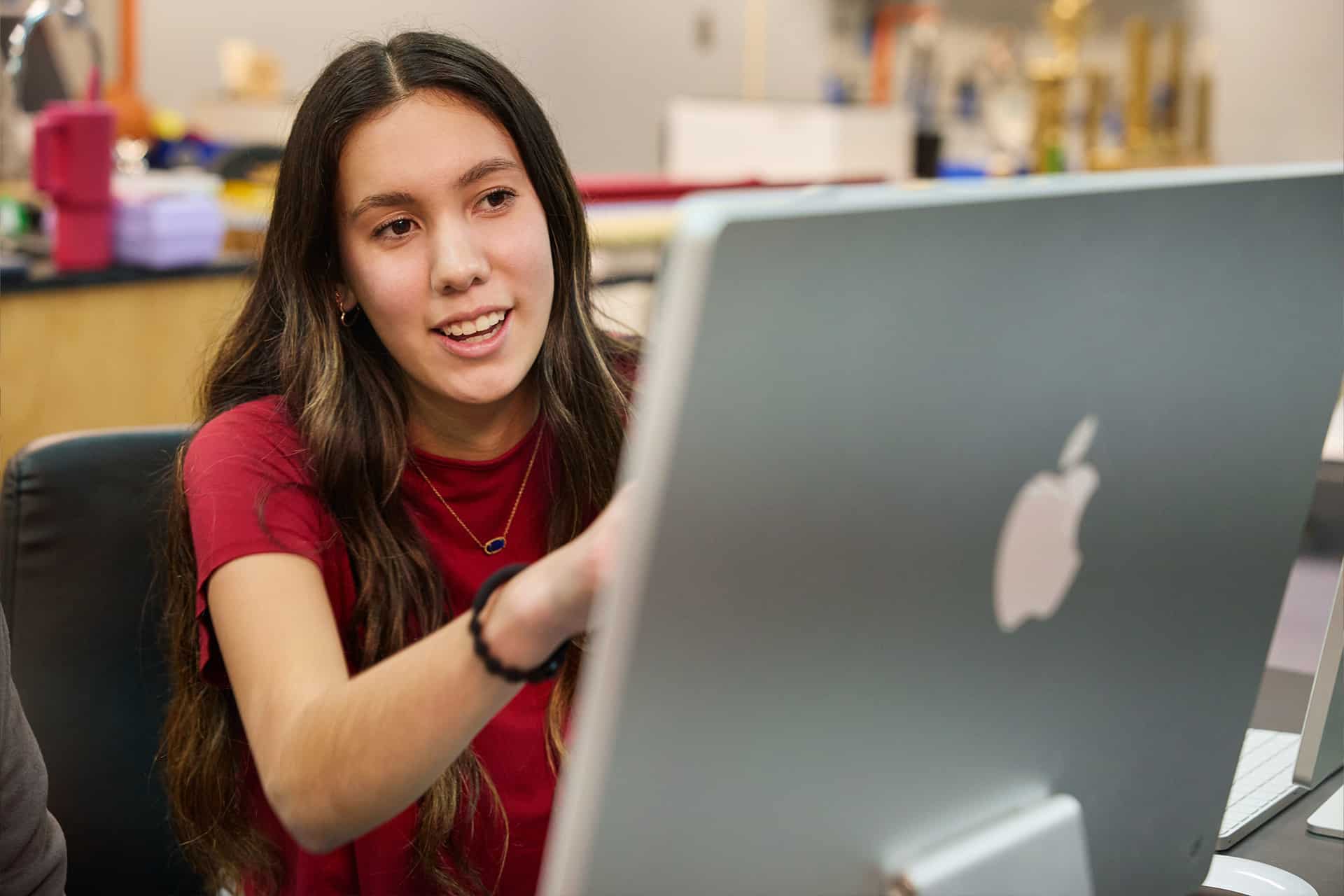 A young woman with long dark hair, wearing a red shirt, sits at a desk and points at a large Apple computer screen displaying an iPad Cherokee language lesson while smiling in a modern classroom or office setting.