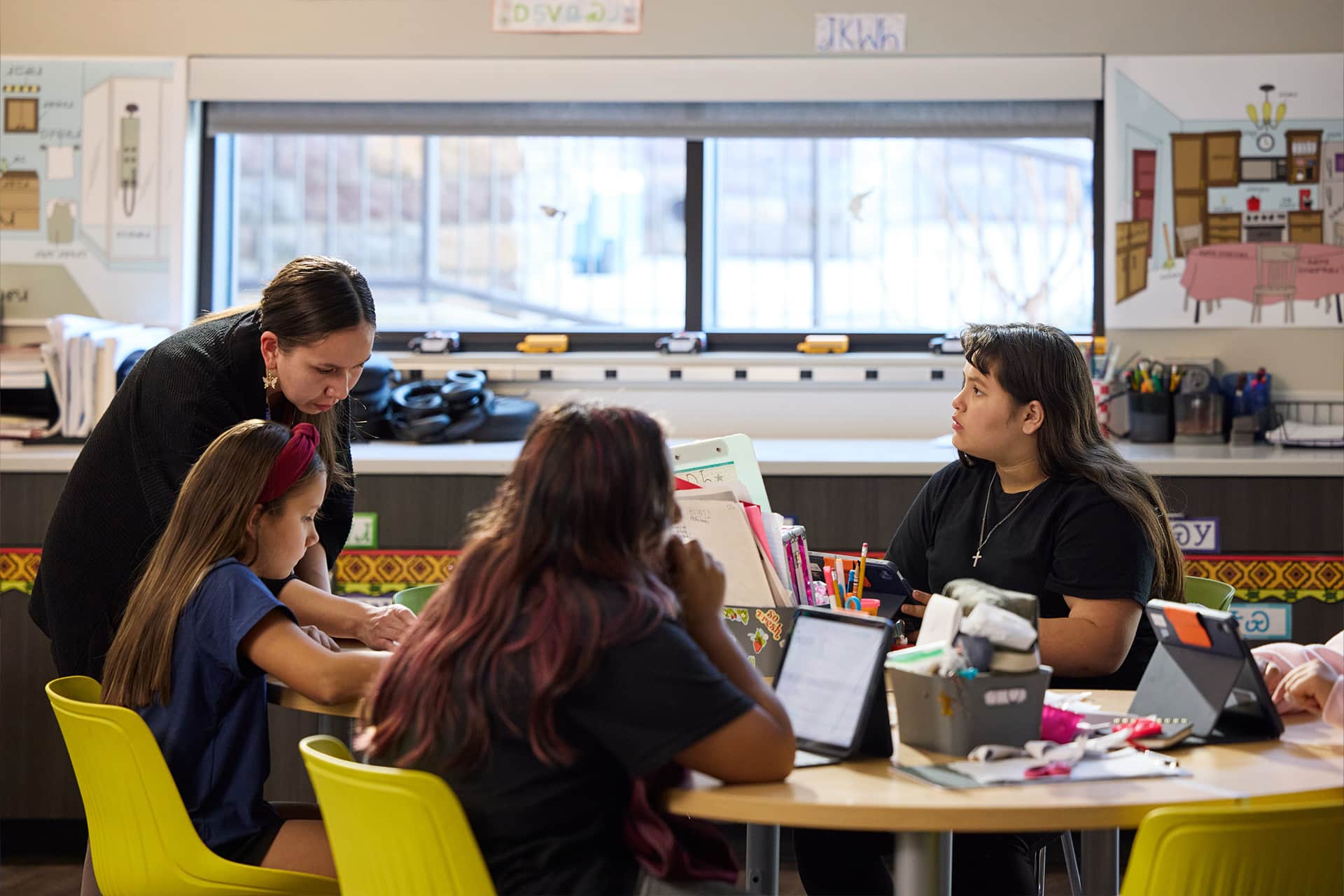 A teacher leans over to help a student at a table, while two other students sit nearby with laptops and an iPad Cherokee in a classroom with bright yellow chairs and educational posters on the wall.