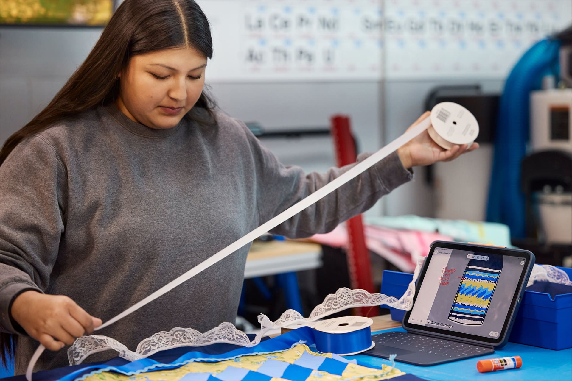 A woman measures white ribbon at a table with sewing supplies, lace, and blue and yellow patterned fabric. An open iPad Cherokee beside her displays a digital design similar to the fabric.