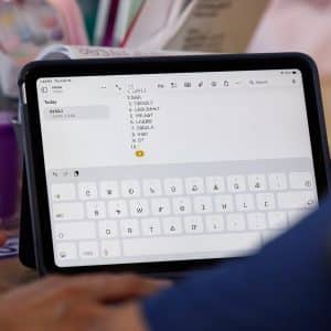 A person uses an iPad Cherokee language app with a digital keyboard displayed, typing a numbered list. Another person’s hand points to the screen. Various items, including a purple cup, are on the table in the background.