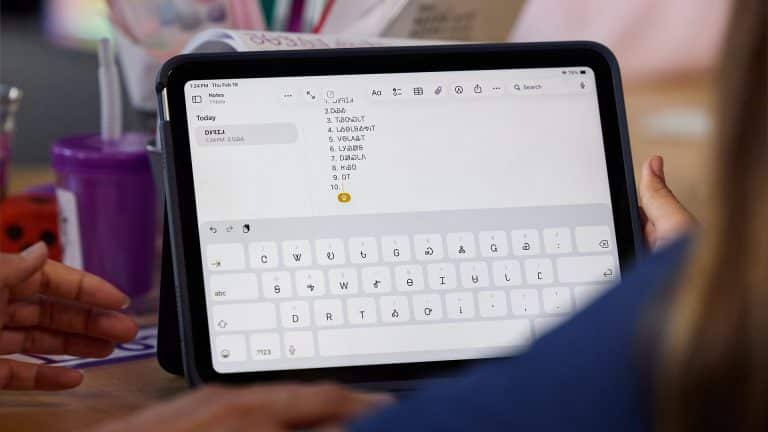 A person uses an iPad Cherokee language app with a digital keyboard displayed, typing a numbered list. Another person’s hand points to the screen. Various items, including a purple cup, are on the table in the background.