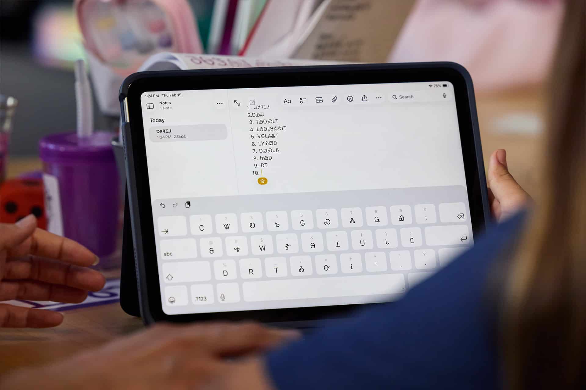 A person uses an iPad Cherokee language app with a digital keyboard displayed, typing a numbered list. Another person’s hand points to the screen. Various items, including a purple cup, are on the table in the background.