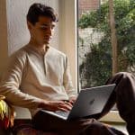 A man sits by a window with greenery outside, working on his laptop during Apple Back to School season. He wears a cream henley shirt and brown pants, relaxing on a colorful blanket in a cozy, well-lit room.