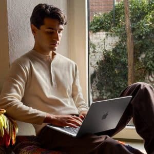 A man sits by a window with greenery outside, working on his laptop during Apple Back to School season. He wears a cream henley shirt and brown pants, relaxing on a colorful blanket in a cozy, well-lit room.